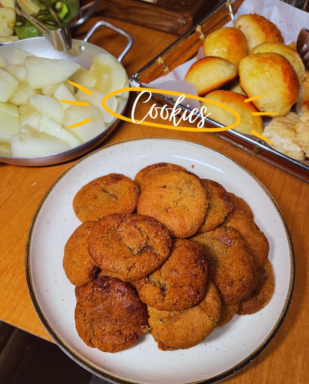 Café colonial: cookies em prato de cerâmica, com pães e frutas ao fundo.