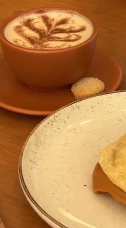 Cappuccino com desenho de folha na xícara de cerâmica, com um biscoitinho no pires.