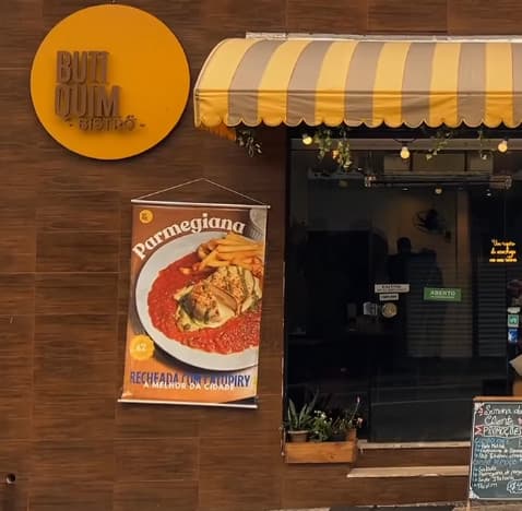 Fachada do Butiquim Bistrô: letreiro redondo amarelo na parede de madeira, toldo listrado amarelo e creme sobre a porta de vidro e plantas na entrada.