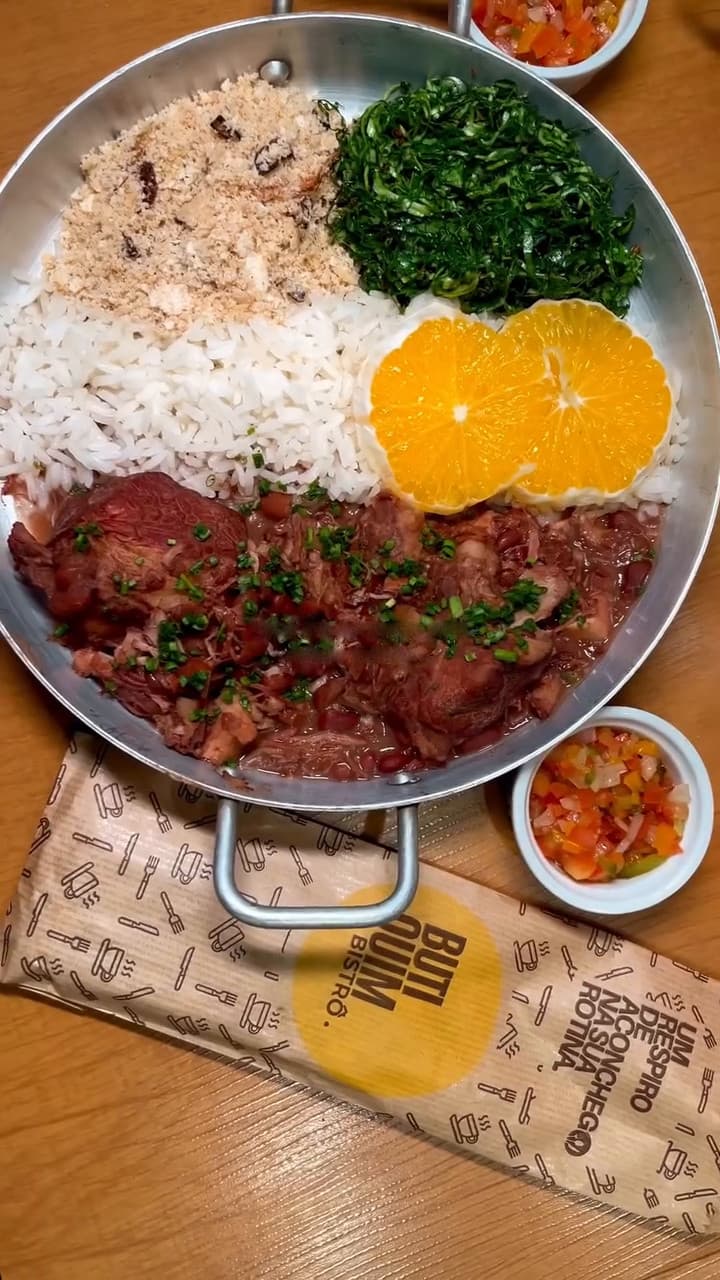 Feijoada do Bistrô na frigideira, com arroz, farofa, couve, rodelas de laranja e vinagrete ao lado, sobre o guardanapo do Butiquim.