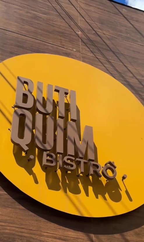 Letreiro redondo amarelo com o nome Butiquim Bistrô em letras marrons, na parede de madeira da fachada.