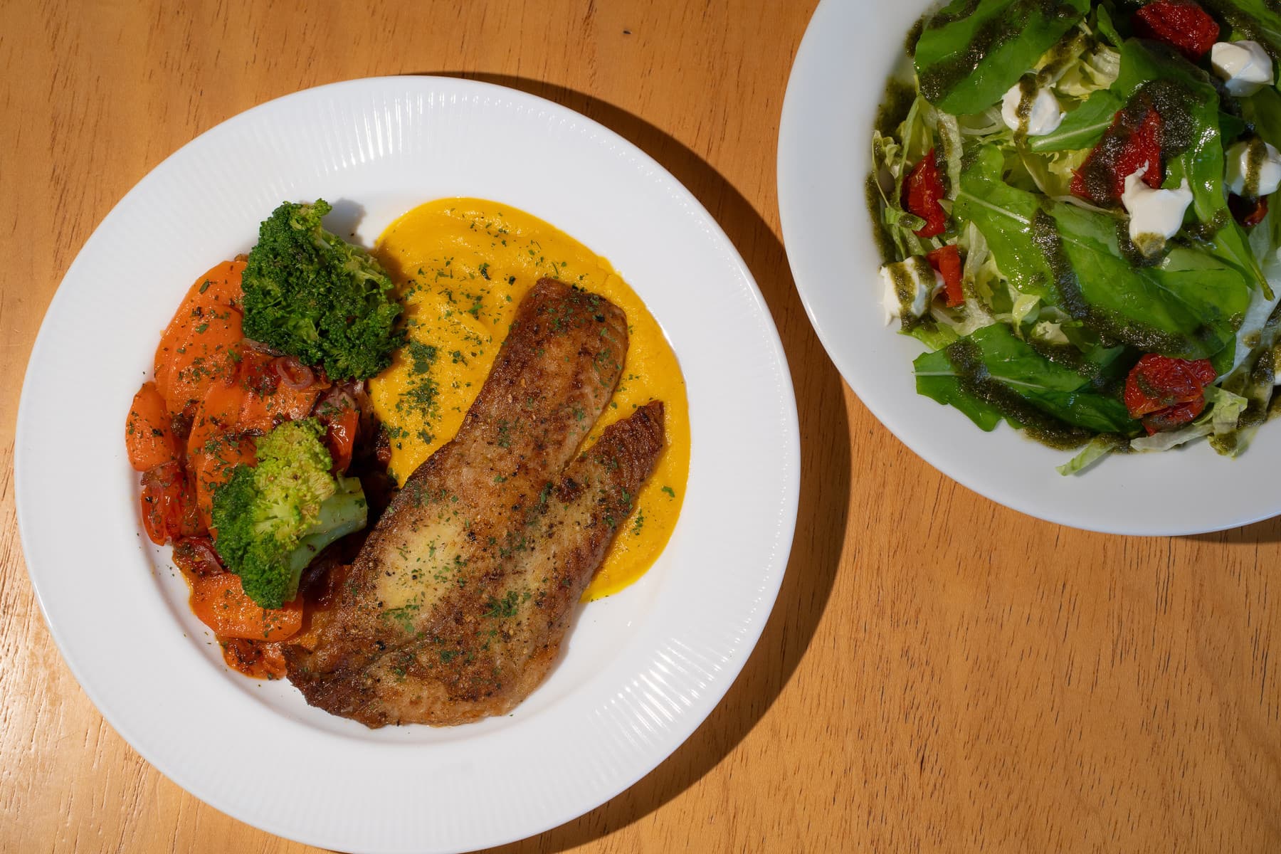 Prato do menu fit com filé de peixe, legumes e purê de abóbora, ao lado de uma salada de folhas com tomate seco e cream cheese.