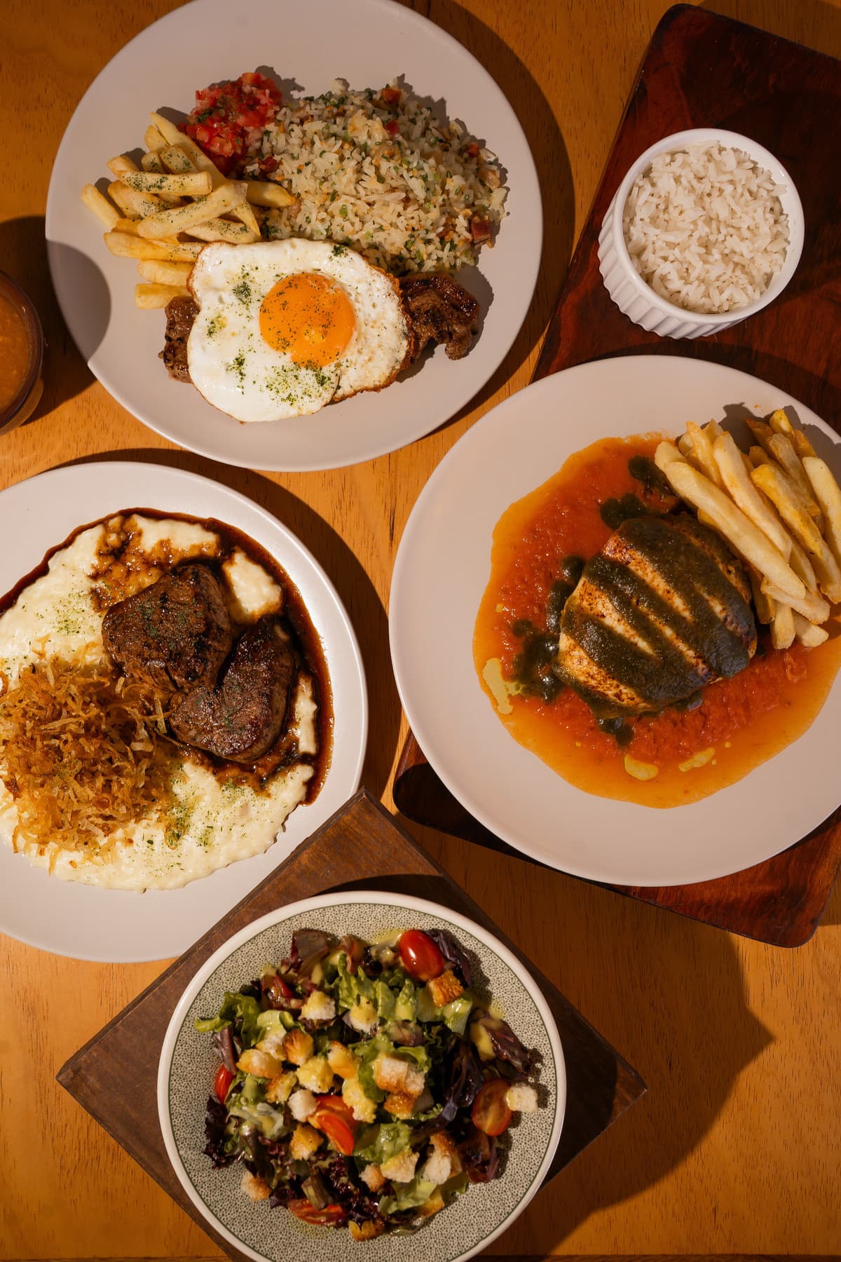 Pratos do almoço vistos de cima sobre mesa de madeira: filé com ovo frito, arroz e fritas; medalhões de filé com arroz cremoso e batata palha; frango com molho de tomate e pesto; e uma salada com croutons.