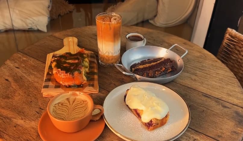 Mesa redonda de madeira com café gelado em camadas, cappuccino, café coado, torrada com pesto na tábua, fatia de bolo de chocolate na frigideira e fatia de bolo com cobertura branca.