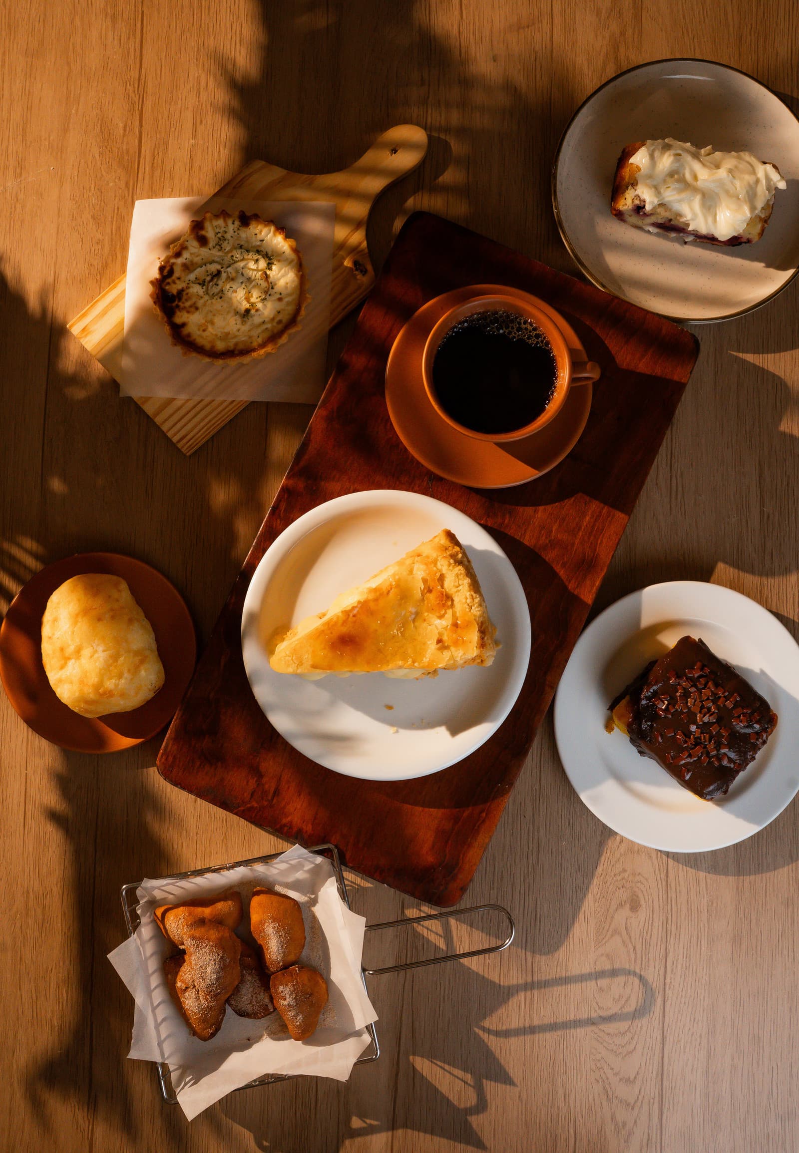 Mesa da cafeteria vista de cima, ao sol: café coado na xícara de cerâmica, quiche, fatia de torta, pão de queijo, bolo de cenoura com brigadeiro, fatia de bolo com cobertura cremosa e bolinhos açucarados na cestinha.