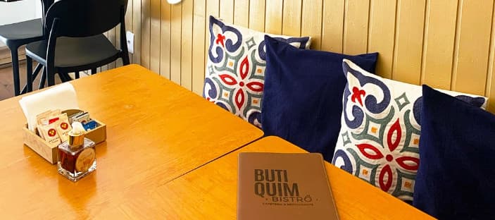 Mesas de madeira junto ao banco com almofadas azul-marinho e estampadas, com o cardápio do Butiquim sobre a mesa.